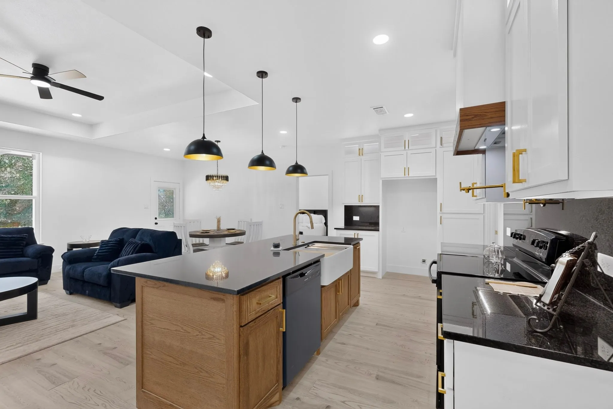 Open-concept kitchen featuring an oversized island with a farmhouse sink, black granite-style countertops, white cabinetry with gold-tone hardware, and light wood-finish flooring