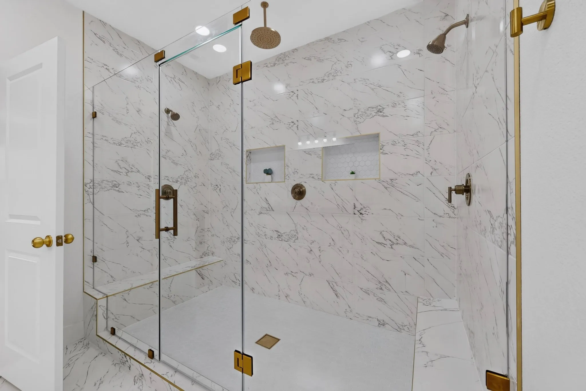 Spacious glass-enclosed shower featuring large format marble-look tile, dual shower heads, integrated bench seating, recessed niches, and brass-finish hardware