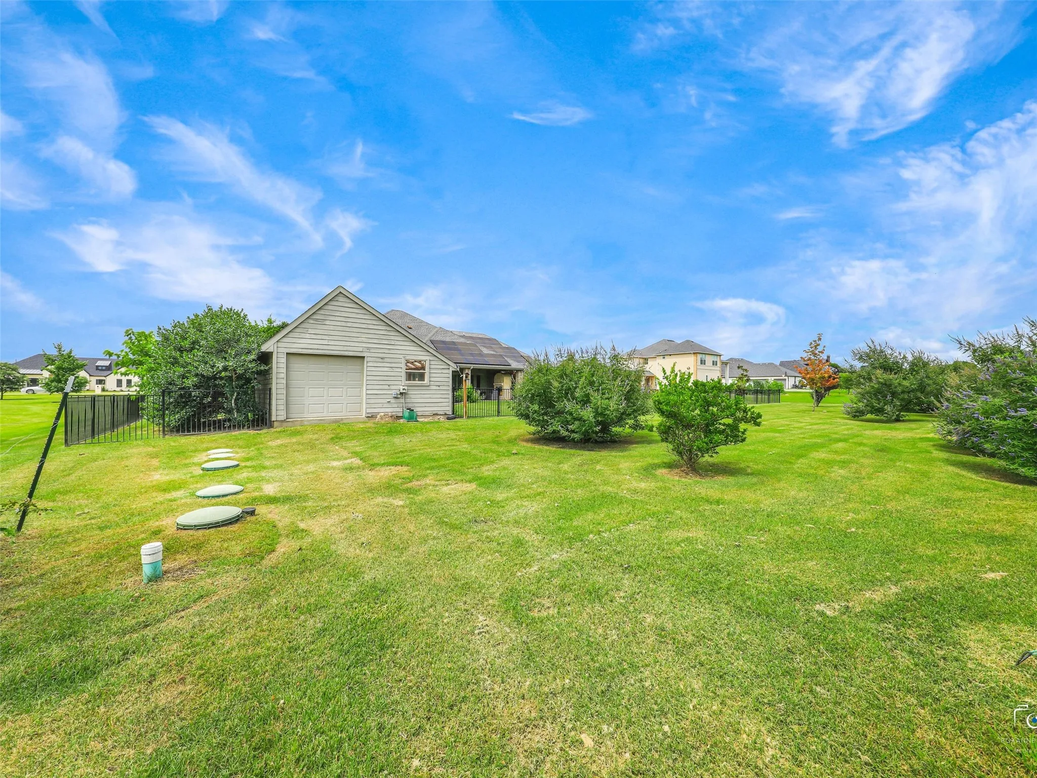 Expansive green lawn featuring a detached garage with a roll-up door, a black metal fence, mature trees, and a stepping stone path