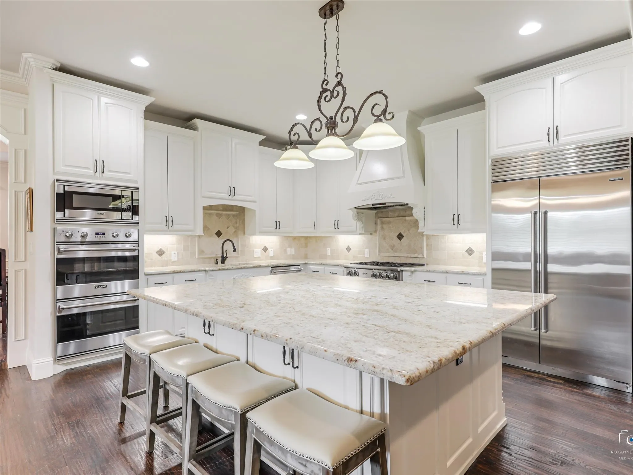 Gourmet kitchen featuring a large central island with light-toned stone countertop, white cabinetry, integrated stainless steel refrigerator, built-in double ovens, and wood-finish flooring