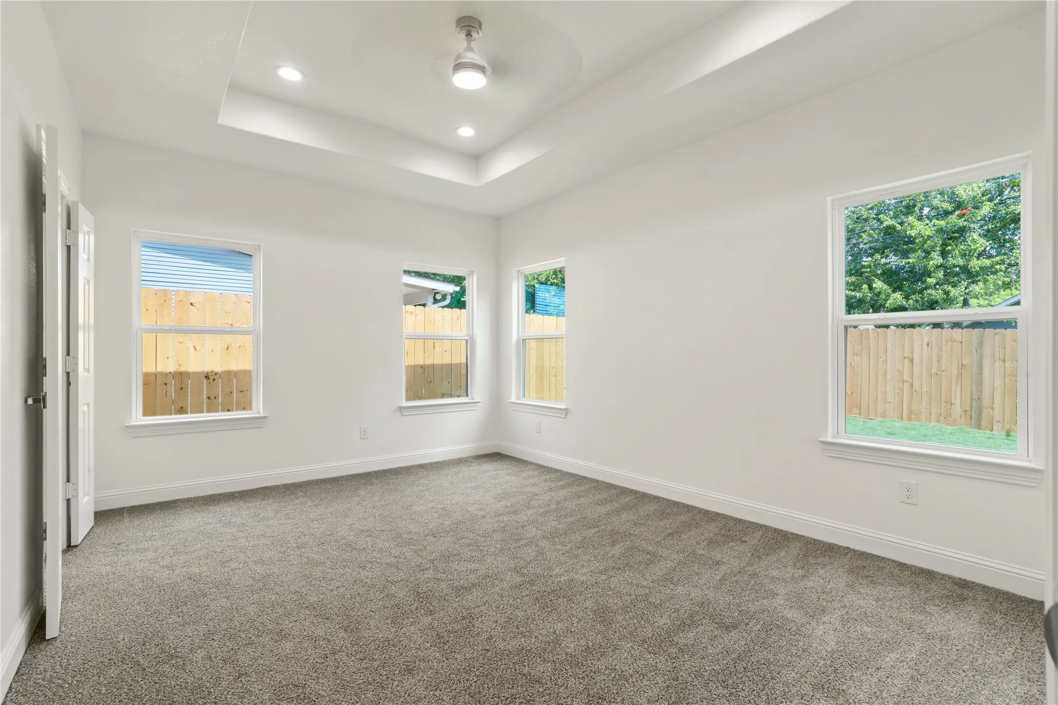 Spacious room featuring a tray ceiling with recessed lighting and a ceiling fan, plush gray carpeting, white walls, and multiple windows