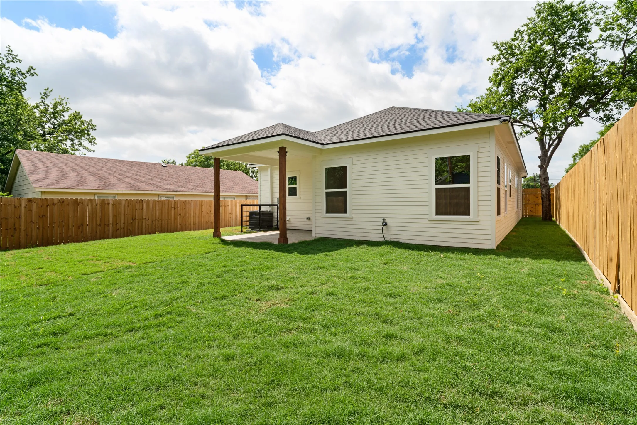 Expansive grassy yard with a covered patio featuring wooden support posts, a dark shingle roof, white siding, and a privacy fence