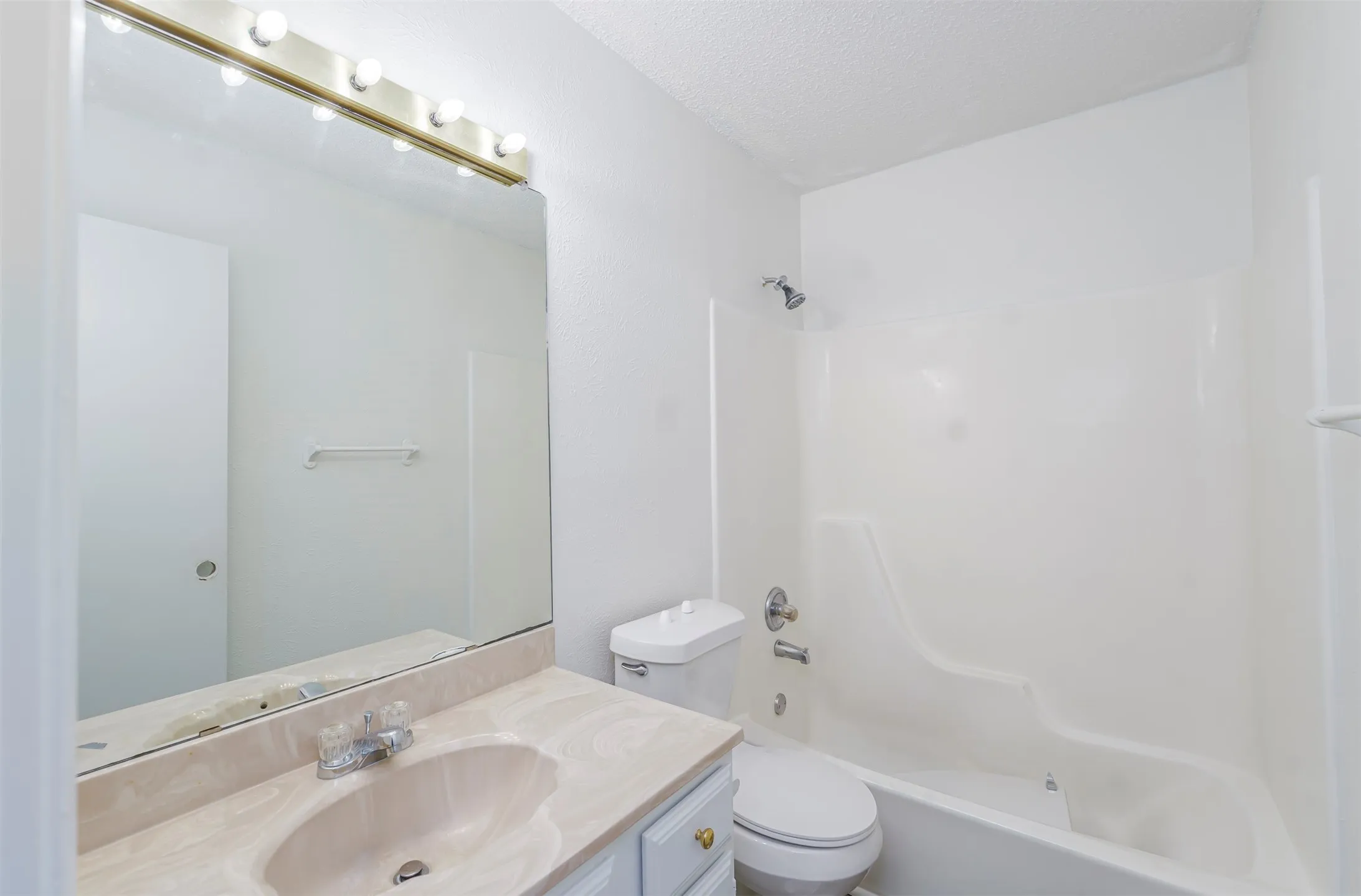 Bathroom featuring a vanity with a drop-in sink and a light-colored countertop, an overhead vanity light with exposed bulbs, a large wall-mounted mirror, a toilet, and a combined shower-tub unit with a chrome showerhead