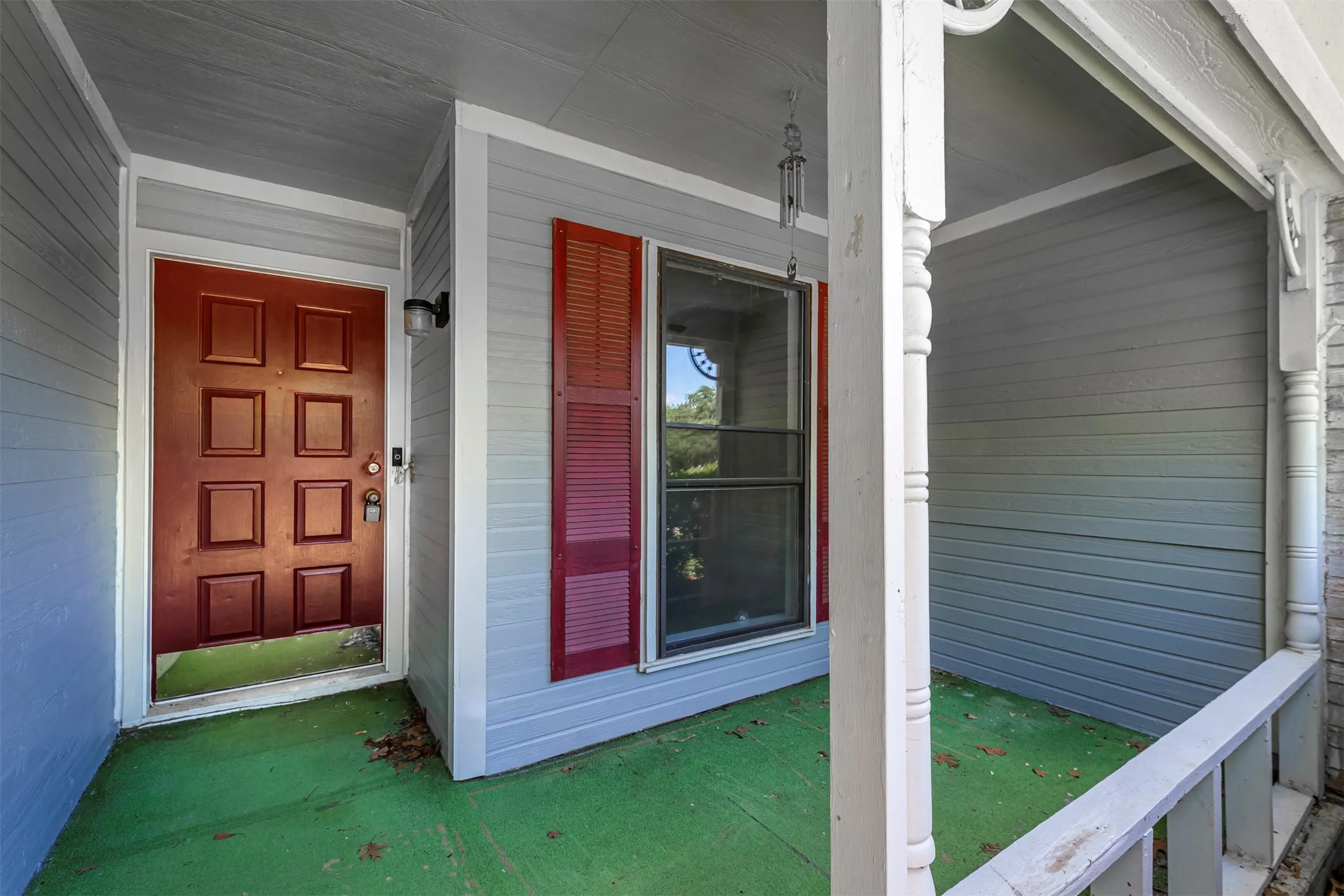 Covered front porch featuring gray siding, a red paneled entry door, a window with red louvered shutters, and white architectural columns
