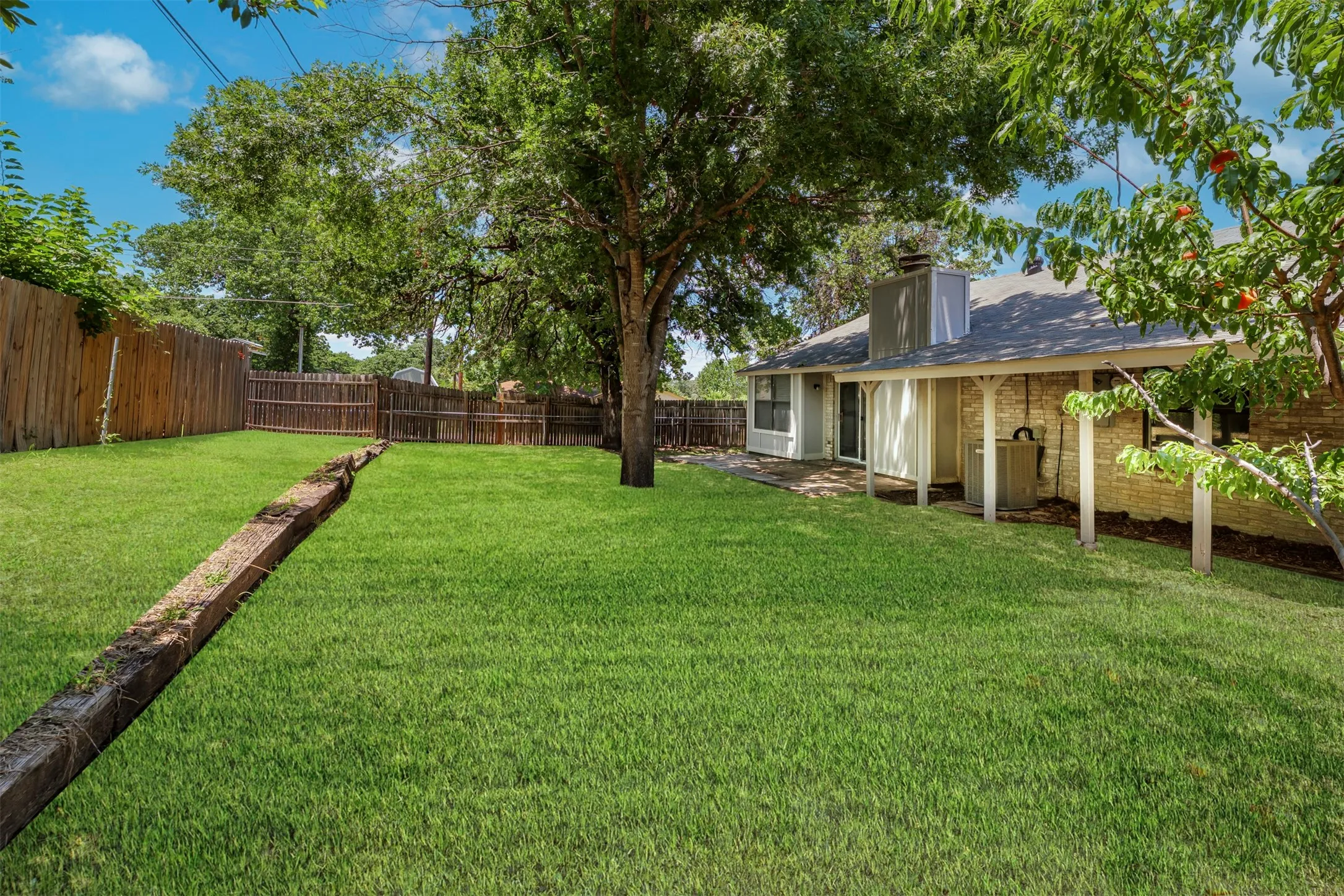 Expansive backyard featuring a manicured lawn, established shade trees, a covered patio with support columns, a brick exterior, and a privacy fence