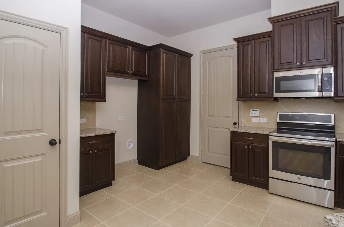 Kitchen featuring rich wood cabinetry, granite countertops, stainless steel appliances, a tiled backsplash, and neutral tile flooring