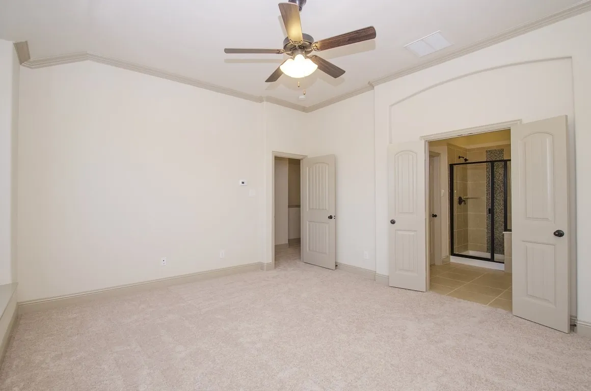 Spacious room featuring neutral carpet flooring, a ceiling fan with integrated lighting, crown molding, and an arched doorway leading to a tiled bathroom with a glass-enclosed shower