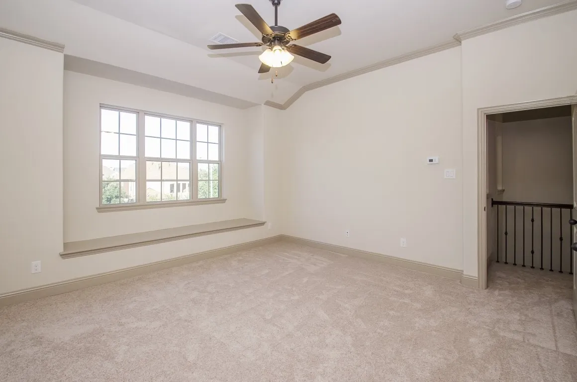 Spacious room featuring vaulted ceilings, a large multi-pane window with a built-in window seat, plush carpet flooring, a ceiling fan with integrated lighting, and crown molding