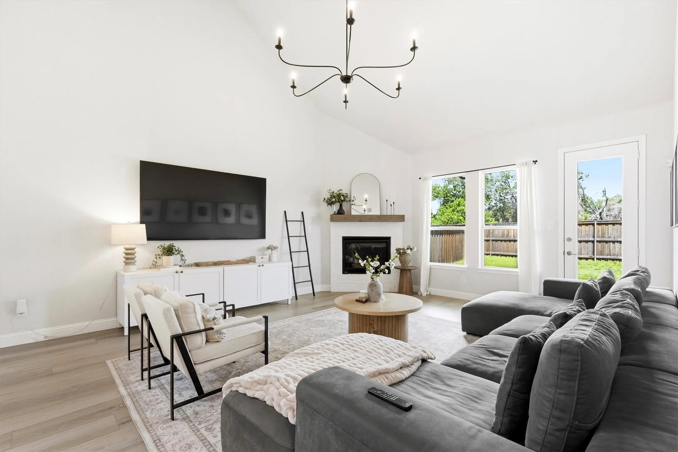 Spacious living area featuring a vaulted ceiling with a modern chandelier, light wood-finish flooring, a white mantel fireplace, and large windows