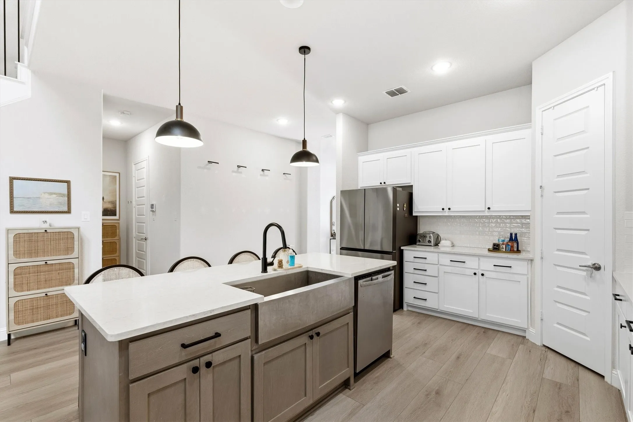 Modern kitchen featuring light wood-finish flooring, white cabinetry with black hardware, stainless steel appliances, and a central island with a farmhouse-style sink and pendant lighting