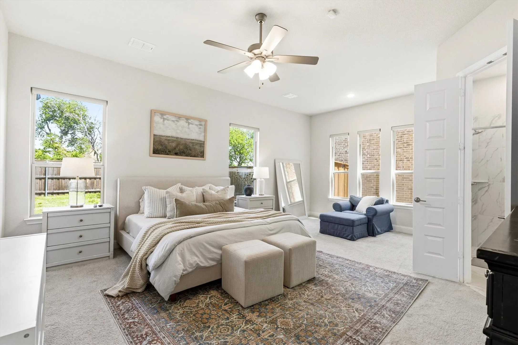 Spacious bedroom featuring light-toned carpeting, multiple windows, a ceiling fan with integrated lighting, and a door opening to a shower with tile surround