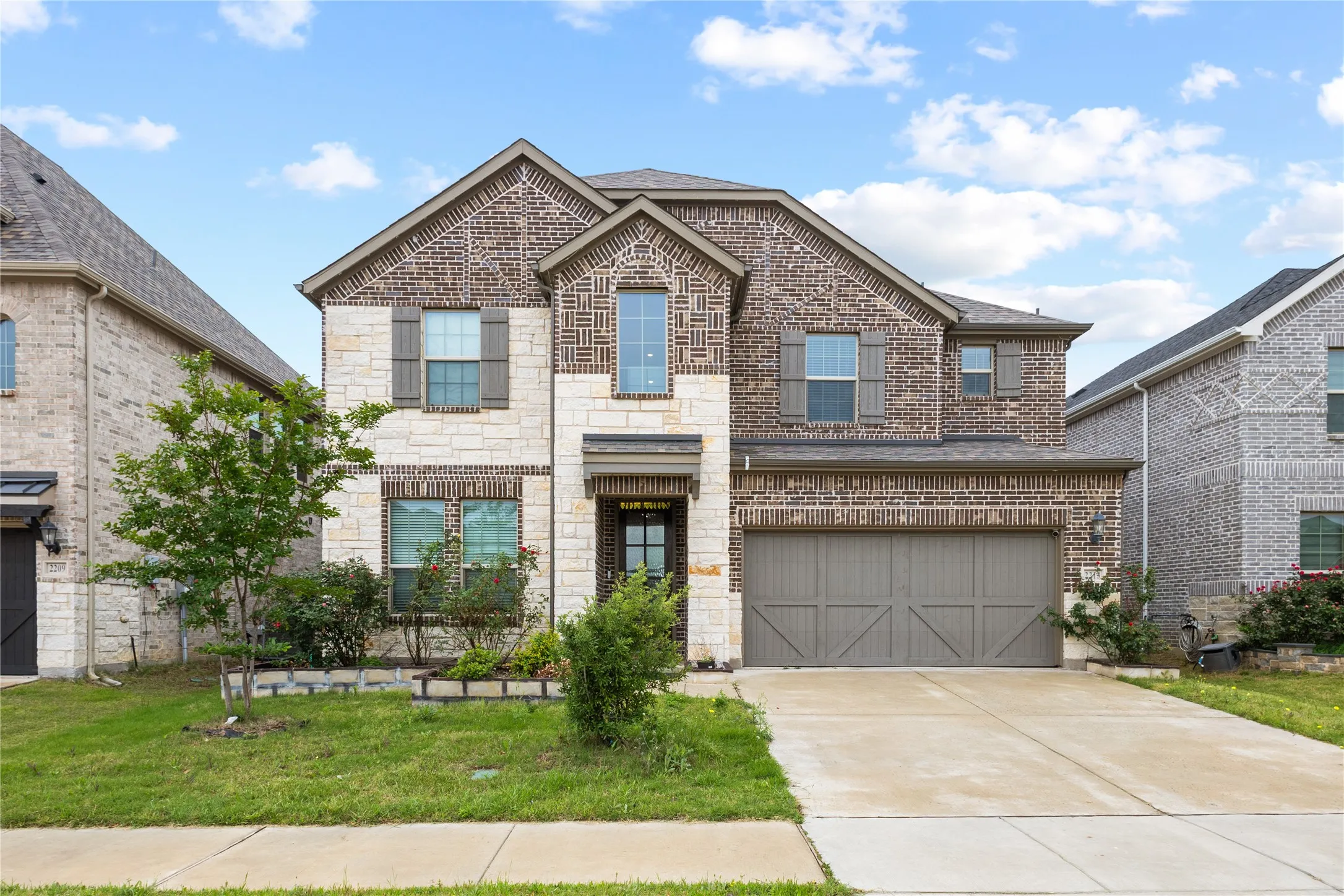 Two-story home featuring a brick and stone facade, contrasting window shutters, an attached two-car garage, concrete driveway, and mature landscaping