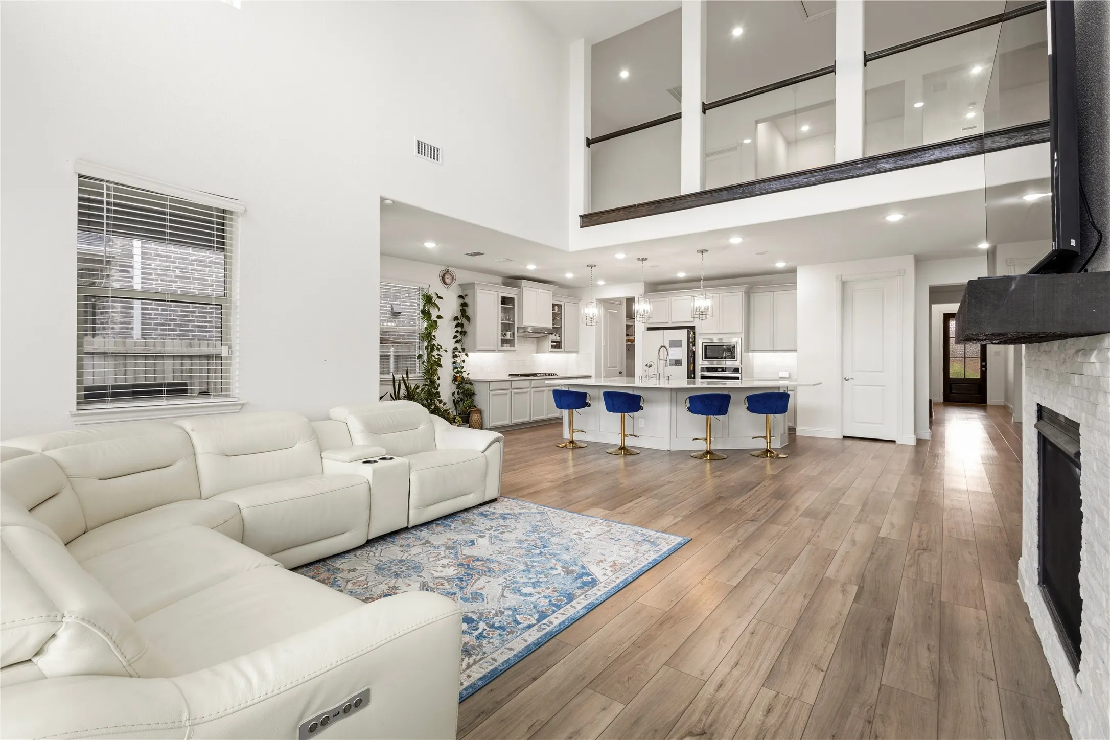 Spacious main living area featuring high ceilings, wood-finish flooring, a white brick fireplace, and an open-concept layout leading to a gourmet kitchen