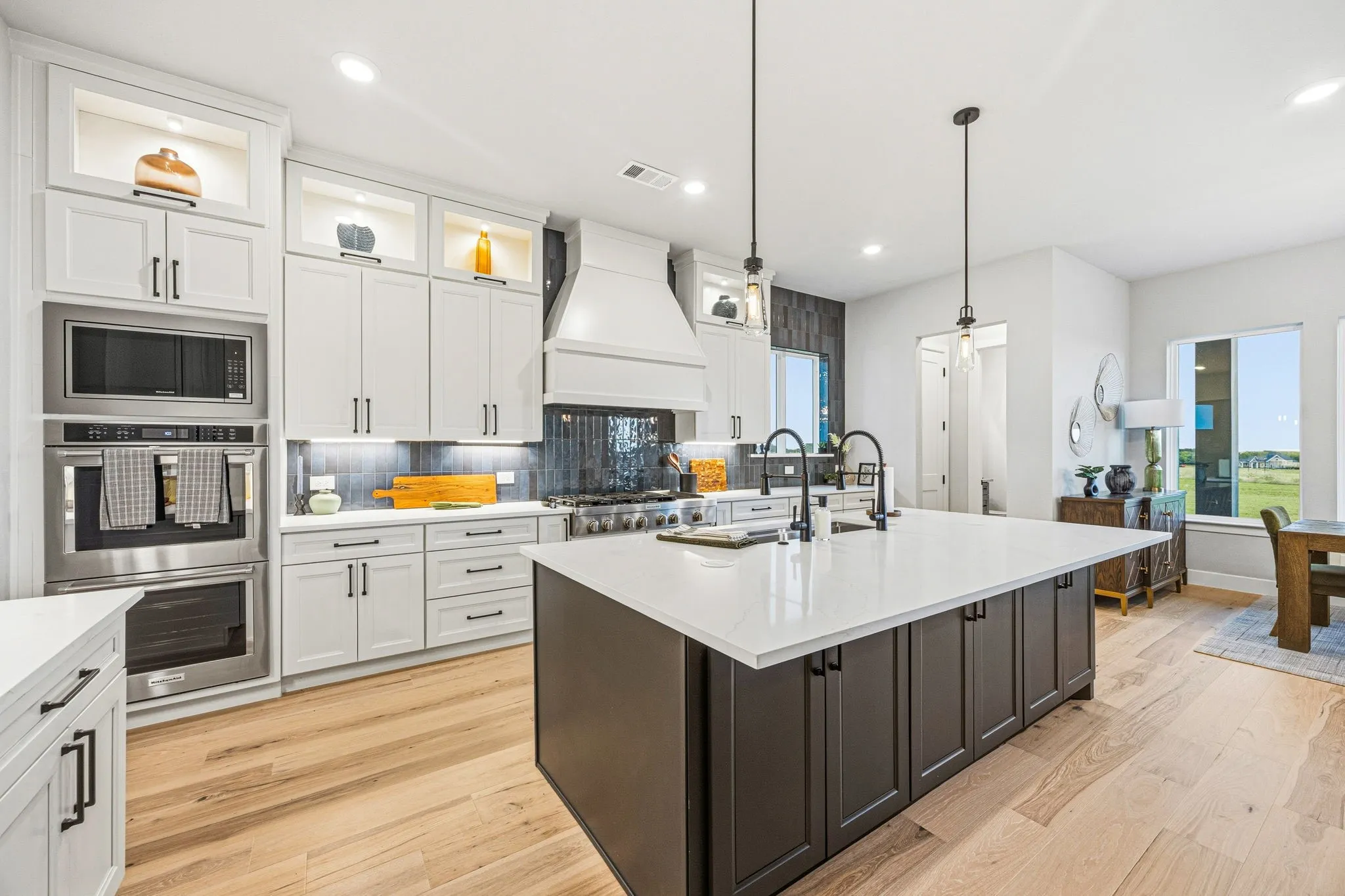Gourmet kitchen featuring a large central island with seating, a built-in oven and microwave, light wood-finish flooring, white upper and lower cabinetry, and a dark accent backsplash