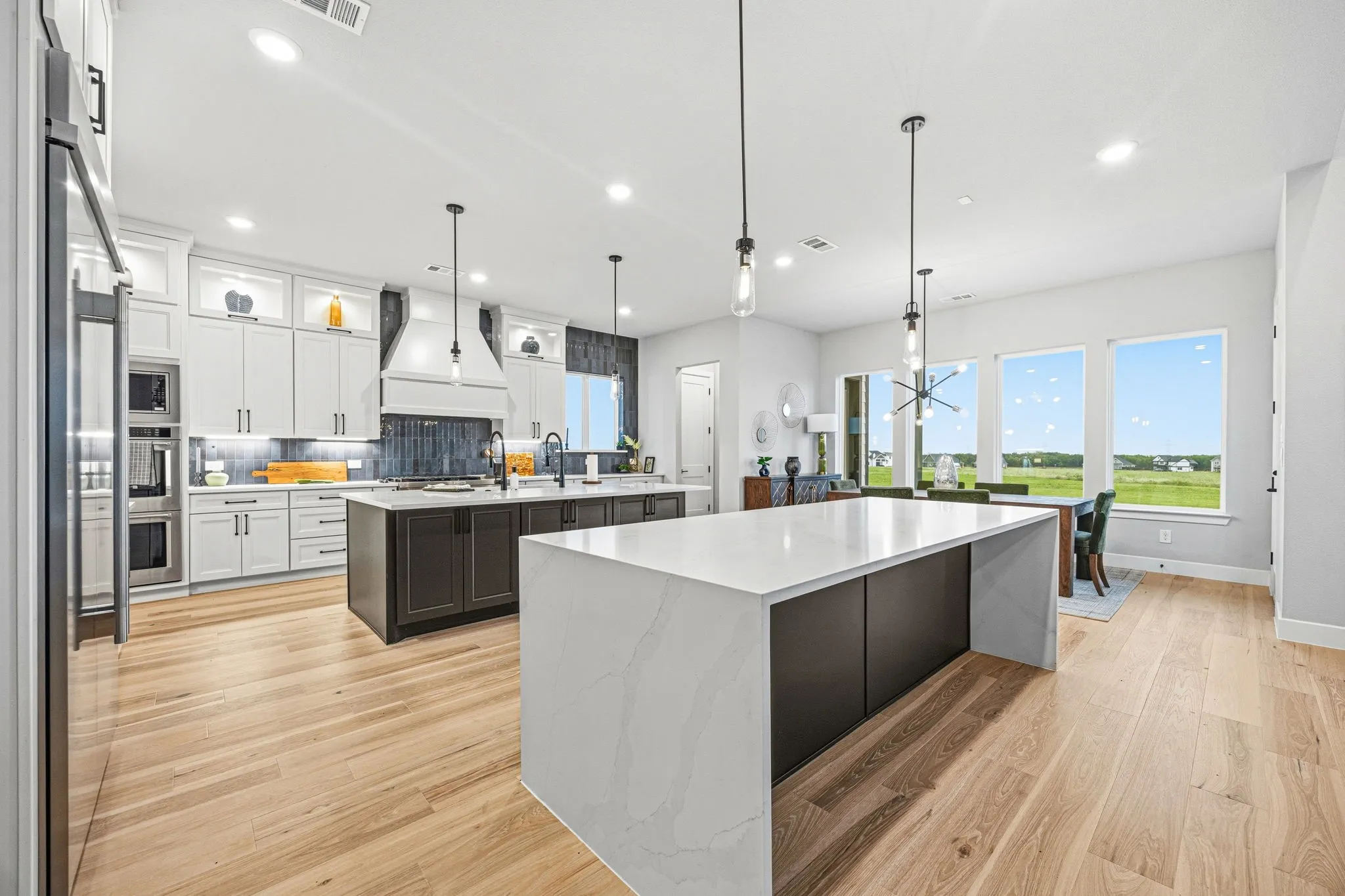 Gourmet kitchen featuring a large Double waterfall island, wood-finish flooring, white cabinetry, integrated stainless steel appliances, and abundant natural light from multiple windows