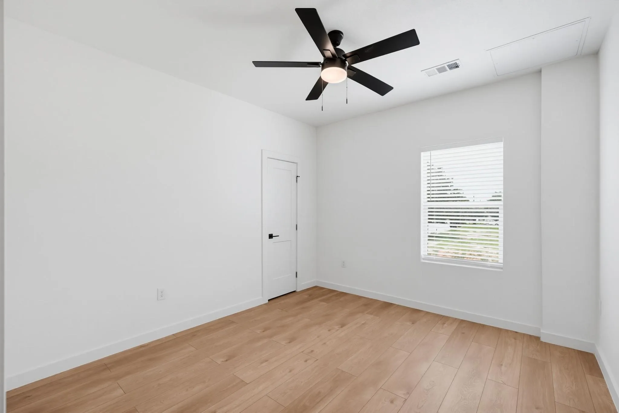 Spacious room featuring wood-finish flooring, a contemporary ceiling fan, crisp white walls, a single window with blinds, and a white door with black hardware