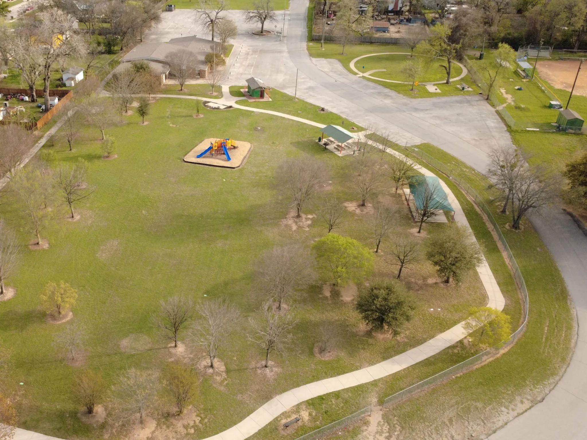 Expansive green space featuring a playground structure, multiple covered picnic areas, a winding paved pathway, and mature trees