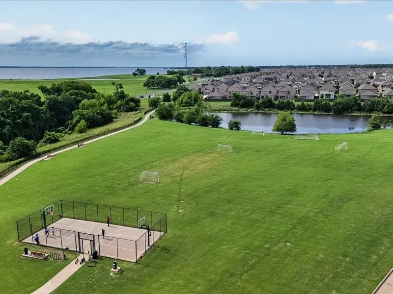 Expansive green space featuring a fenced basketball court, multiple soccer goals, a paved walking path, and a serene body of water