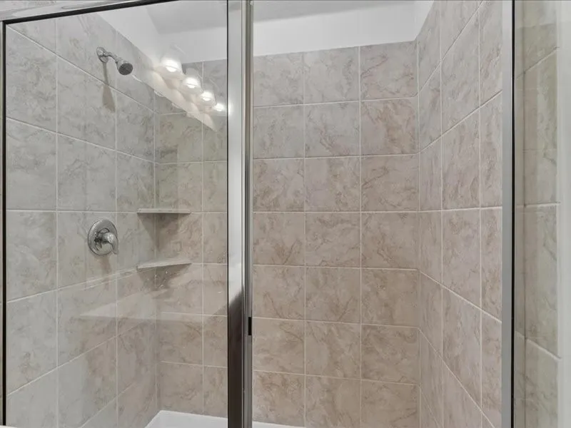 Glass-enclosed shower featuring light-toned tilework, built-in corner shelving, a chrome showerhead, and a matching lever-style faucet