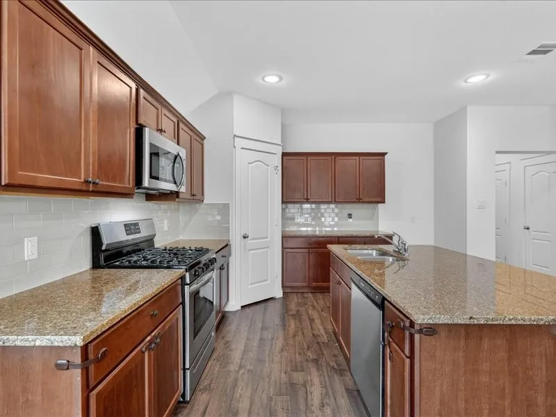Kitchen featuring wood-finish cabinetry, granite-style countertops, stainless steel appliances, a tile backsplash, and wood-finish flooring