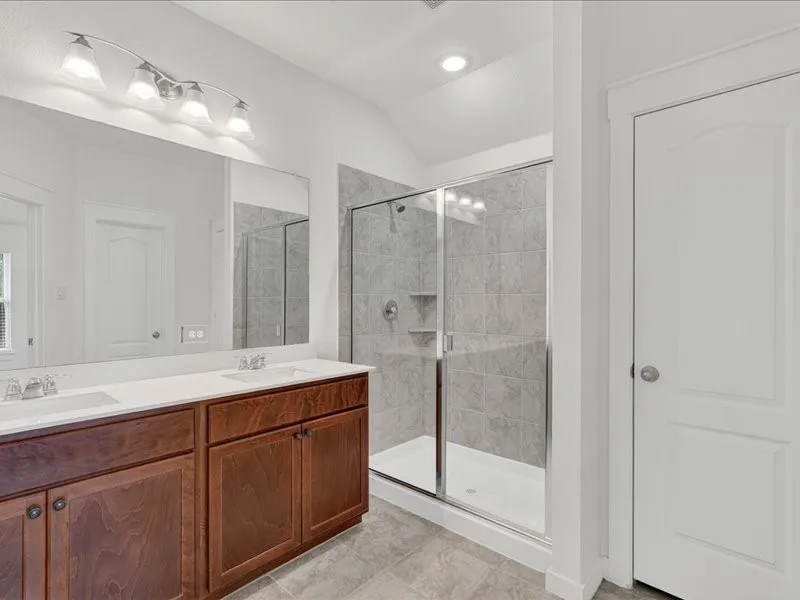 Bathroom featuring a dual-sink vanity with wood cabinetry and white countertops, a large frameless mirror, and a glass-enclosed shower with tiled walls and built-in shelving