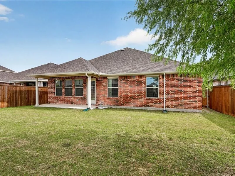 Expansive backyard featuring a covered patio, red brick exterior, multiple windows with grilles, and a privacy fence