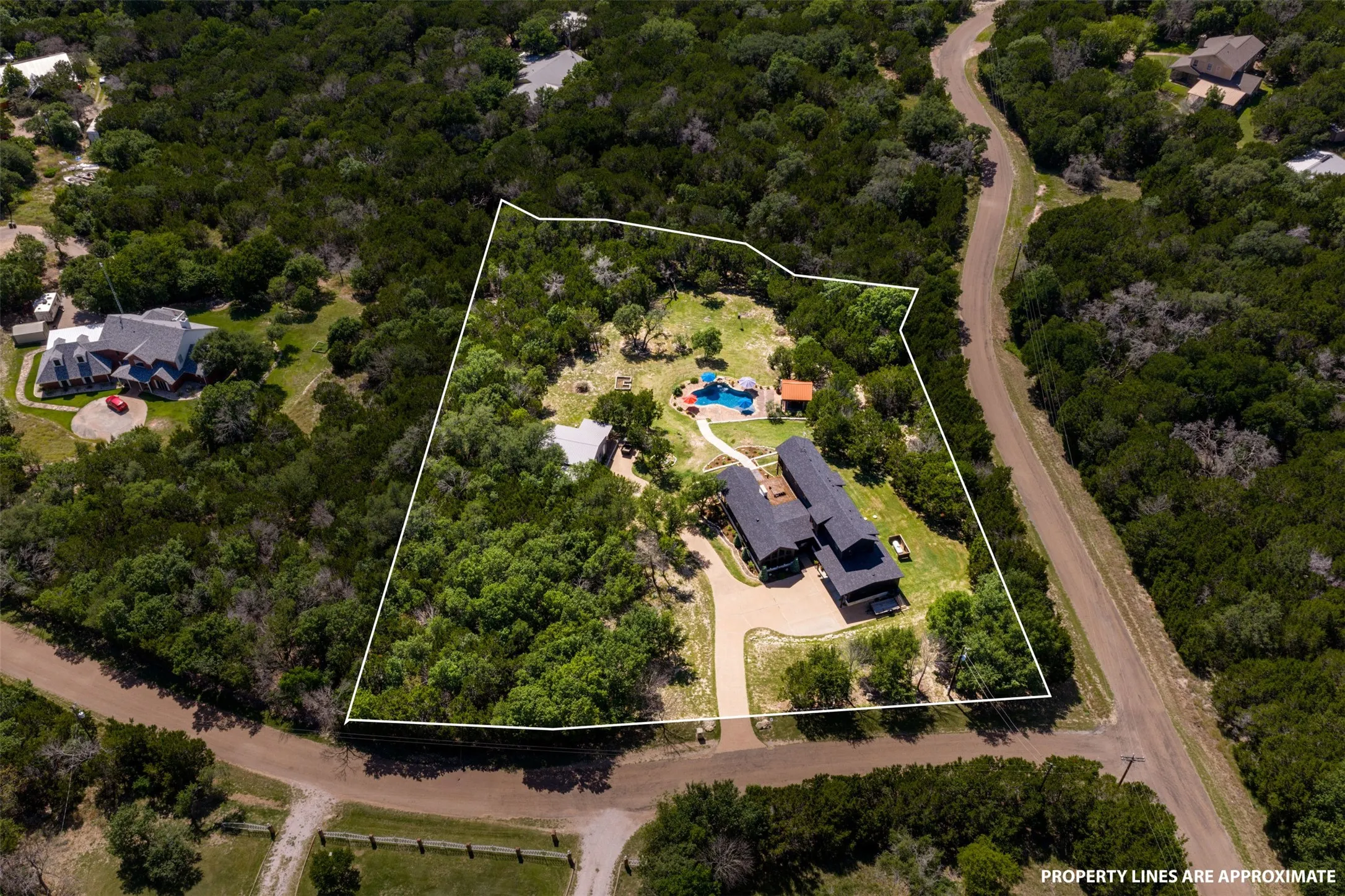 Expansive property featuring a dark-toned residential structure, a detached guest house, a visible swimming pool, a driveway, and extensive surrounding tree cover