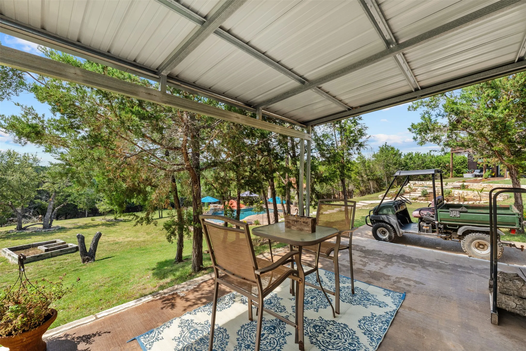 Covered patio off shop with metal roofing and structural beams, overlooking a verdant landscape with mature trees and a distant pool area