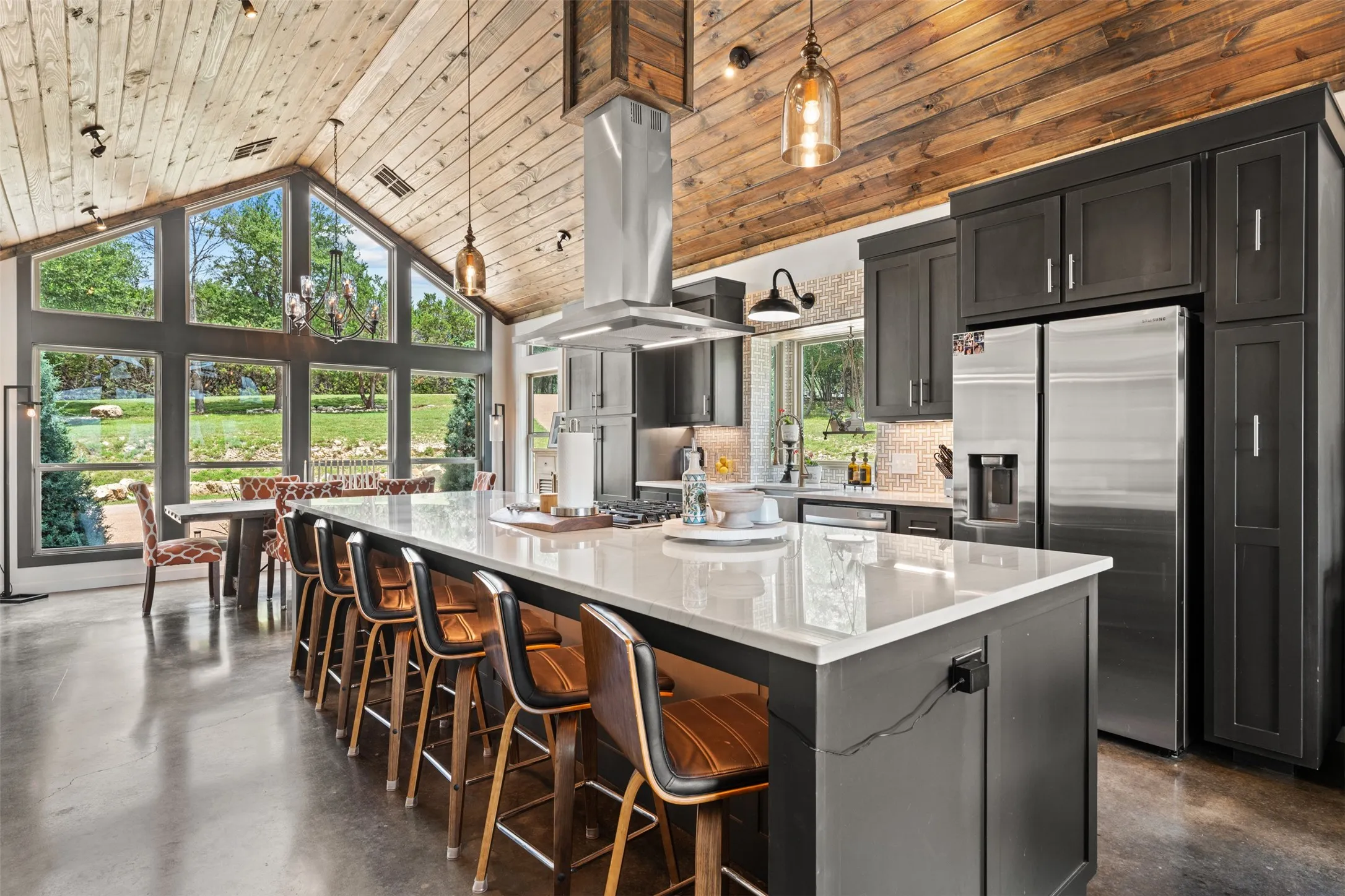 Vaulted wood-paneled ceiling kitchen featuring an expansive central island with a light-toned countertop, stainless steel appliances, and dark cabinetry
