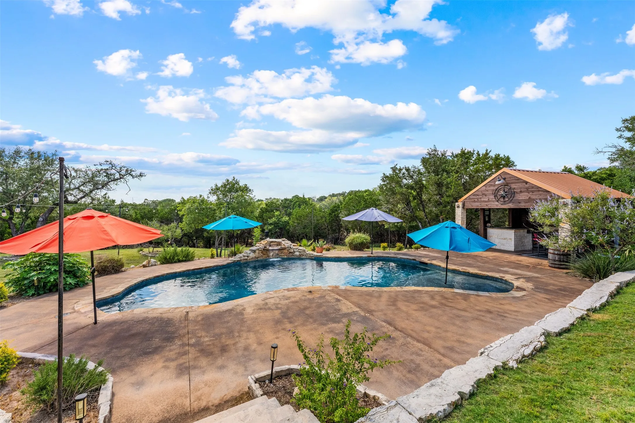 Freeform swimming pool with rock waterfall feature, expansive deck, and a stone and wood-clad outdoor bar structure