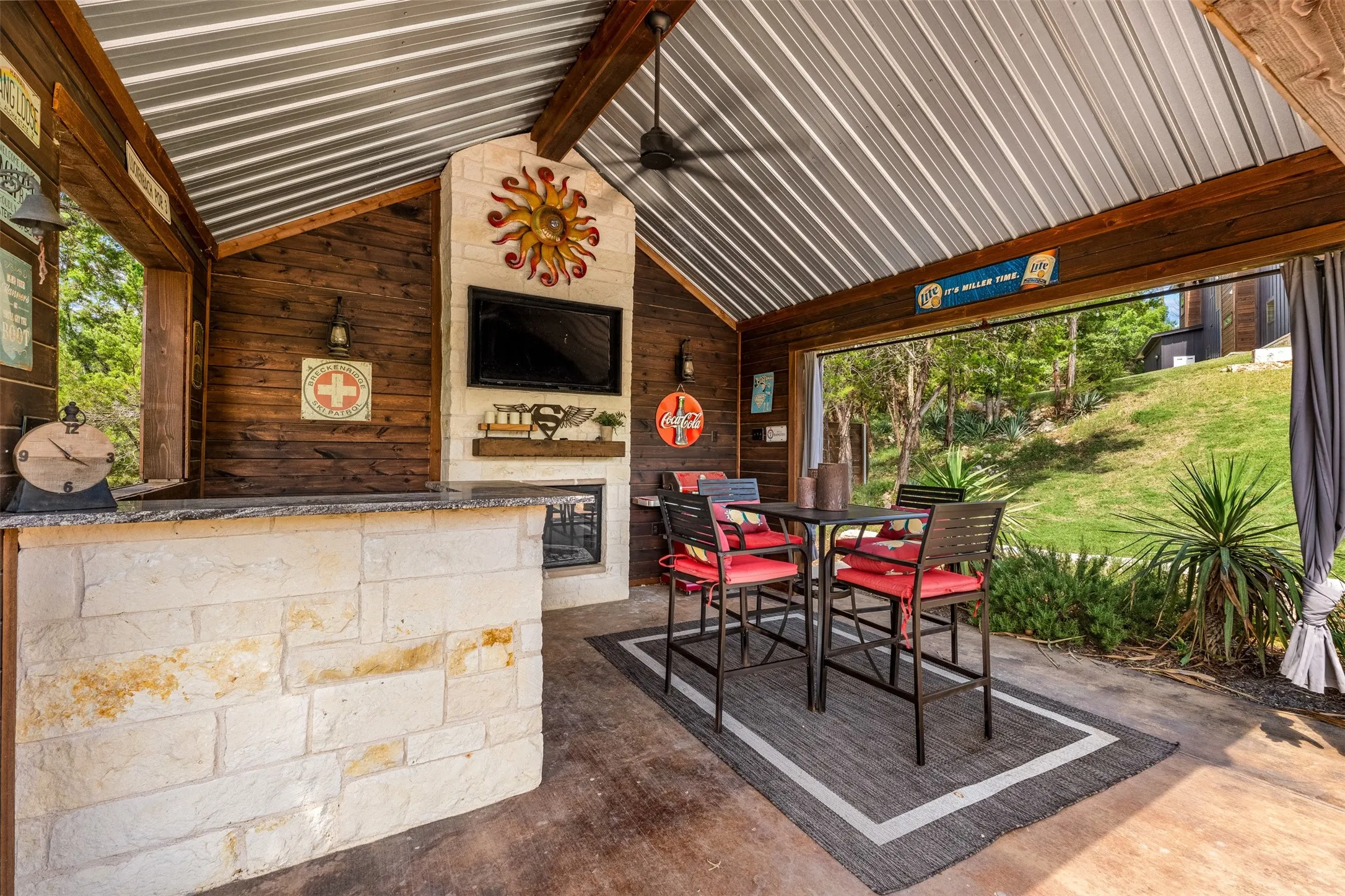 Covered outdoor entertainment area featuring a stone-clad bar with a countertop, a stone fireplace, and a wood-paneled accent wall