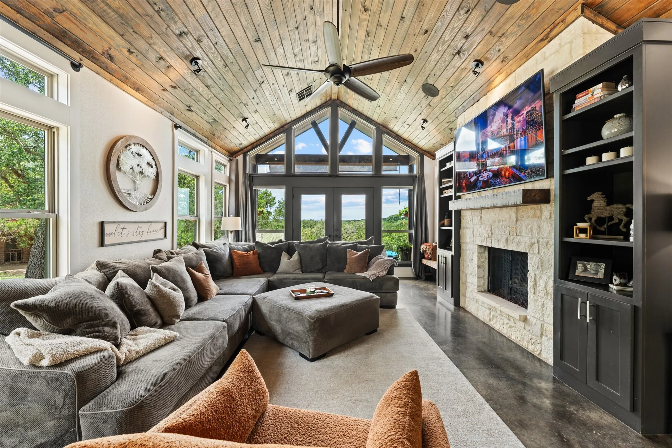 Vaulted wood-plank ceiling with exposed beams, stone fireplace, built-in shelving, and expansive windows