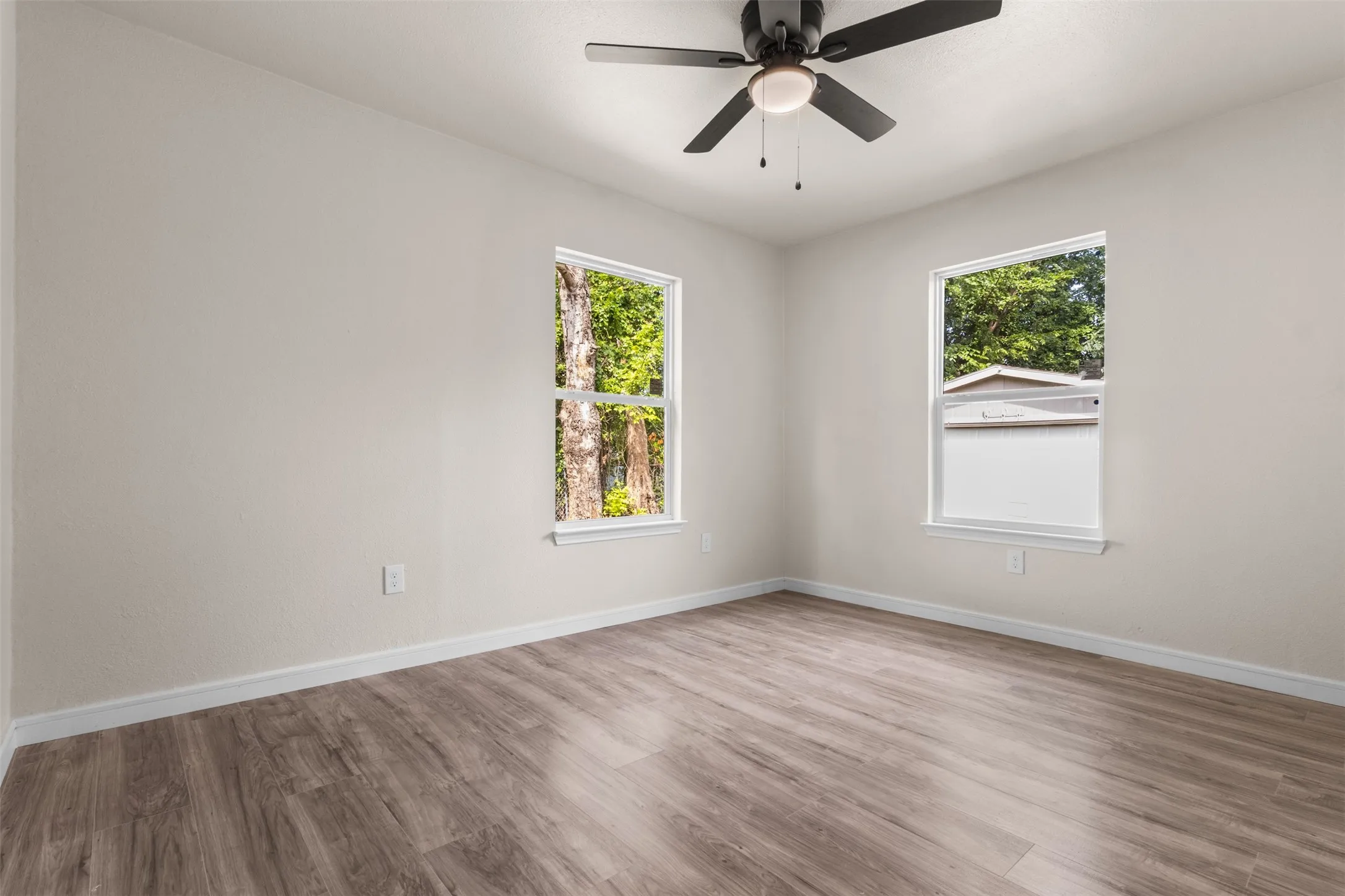 Spacious room featuring wood-finish flooring, light neutral wall paint, two windows with white trim, and a ceiling fan with integrated lighting
