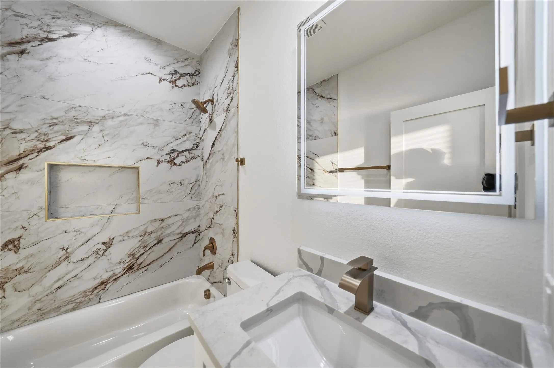 Bathroom featuring a white bathtub with gold-finish fixtures, a built-in shower niche, and full-height marble-patterned wall tiling