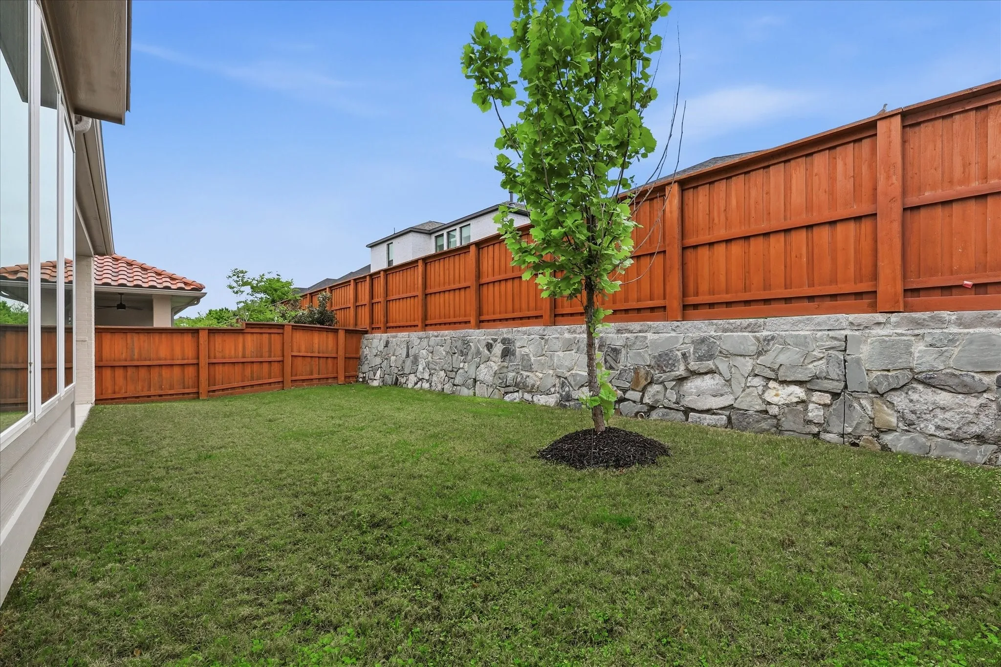 Fenced backyard featuring a manicured lawn, a mature tree, and a stone retaining wall