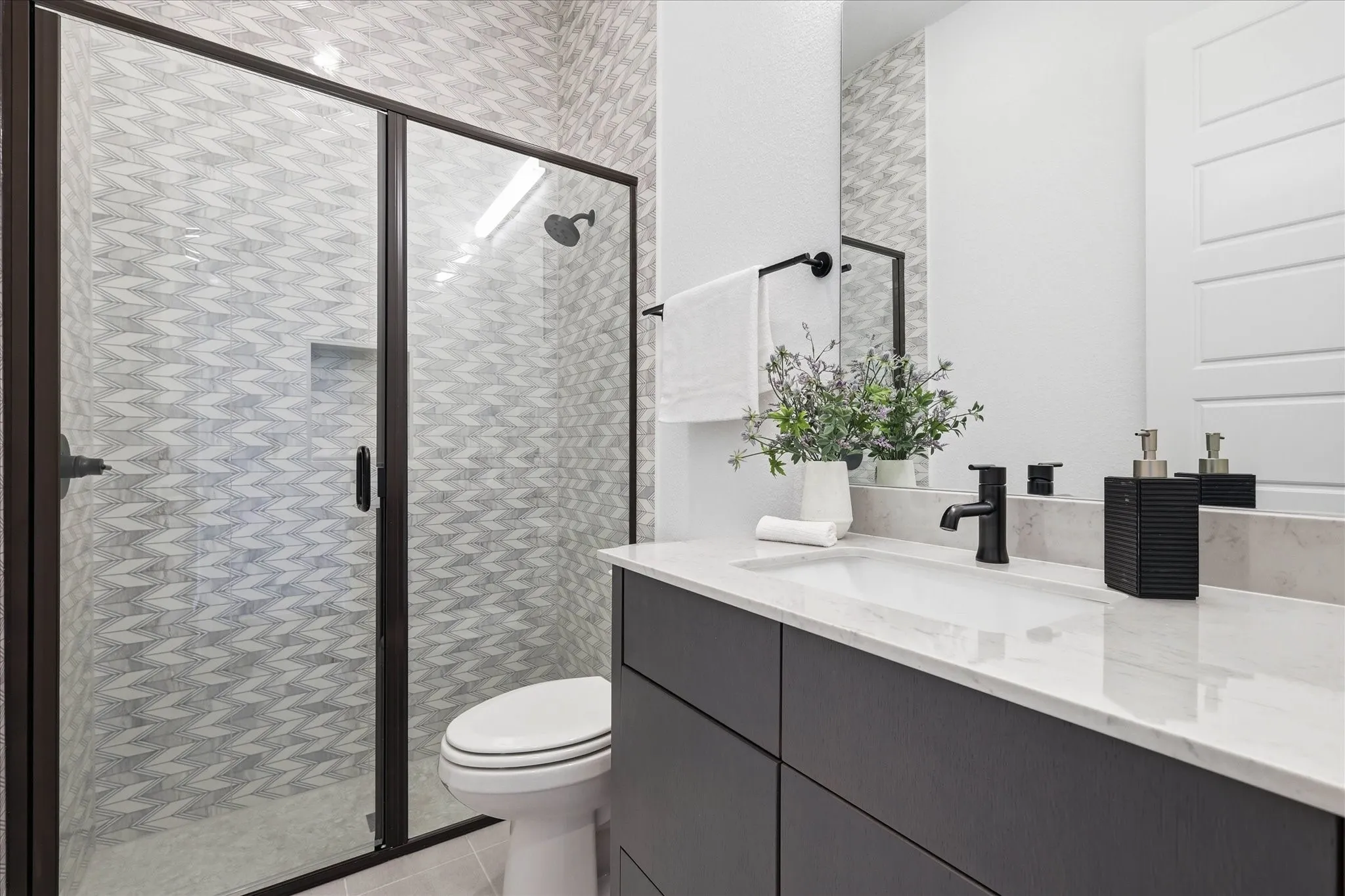 Modern bathroom featuring a frameless glass shower enclosure with dark bronze accents, patterned tile shower surround, single vanity with a white countertop, dark flat-panel cabinetry, and matte black plumbing fixtures