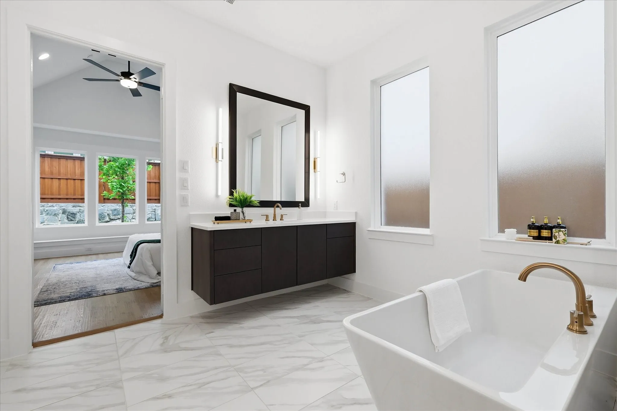 Modern bathroom featuring a rectangular freestanding tub with a brass-finish faucet, dual tall frosted windows, and white marble-look tile flooring