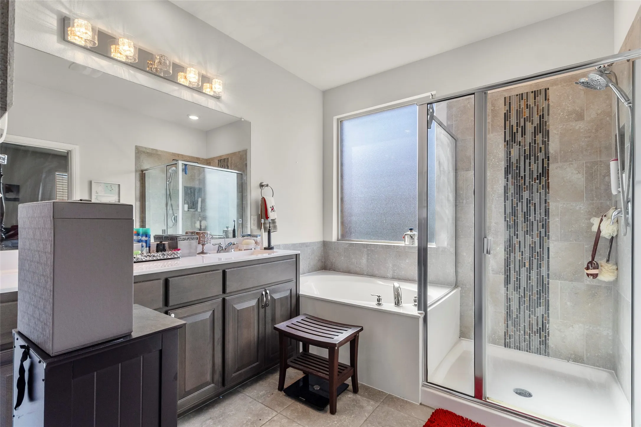 Bathroom featuring a glass-enclosed shower with decorative tile accent, a built-in soaking tub, a large vanity with stone-finish countertop, and extensive mirror