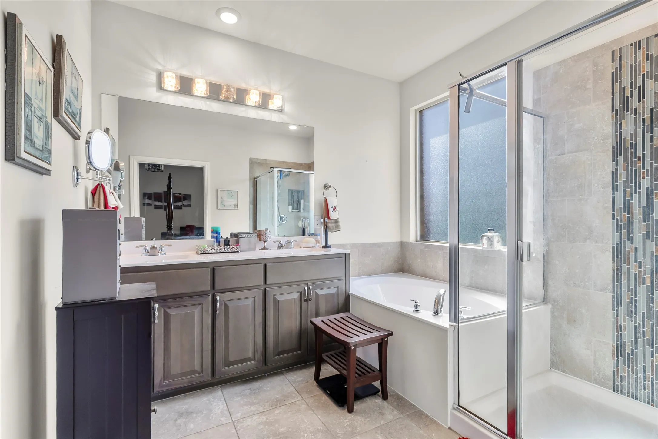 Bathroom featuring a dual vanity with undermount sinks, a large mirror, a separate soaking tub, a glass-enclosed shower with tiled walls and a mosaic accent, and tiled flooring