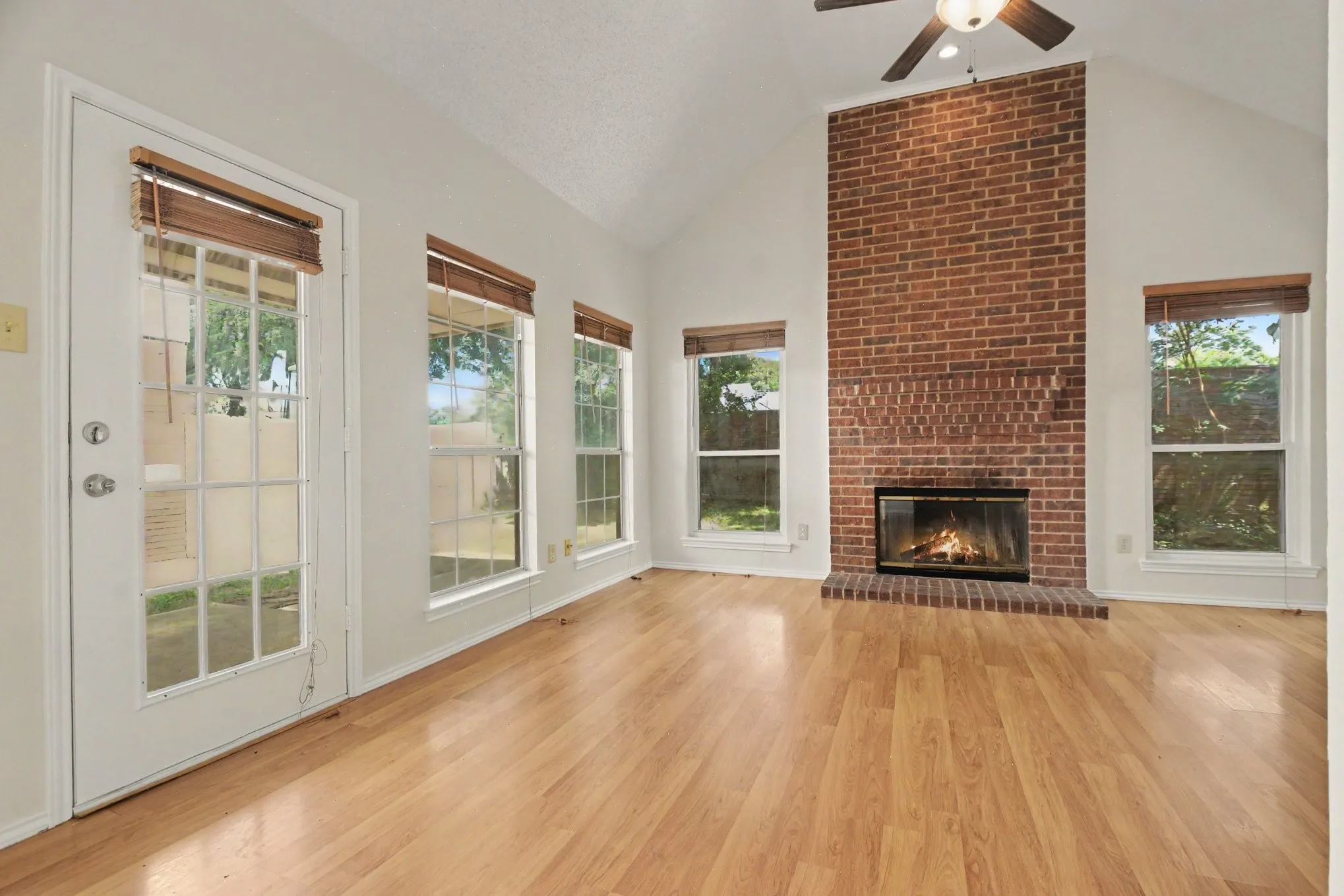 Vaulted ceiling living area featuring a floor-to-ceiling brick fireplace, wood-finish flooring, and multiple windows with wooden blinds