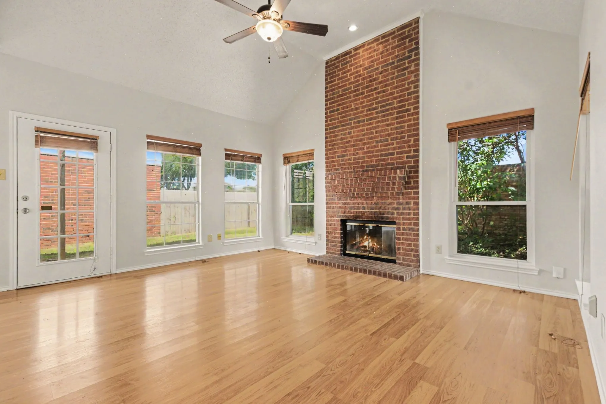 Spacious living area featuring a vaulted ceiling with a ceiling fan, a full-height brick fireplace, wood-finish flooring, and multiple windows providing ambient light