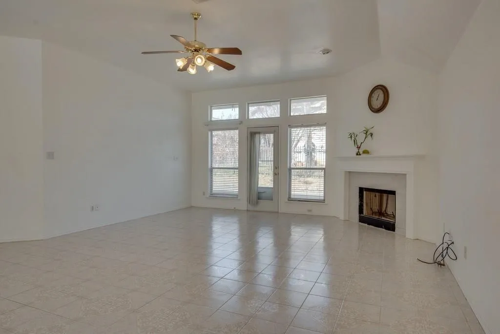 Spacious room featuring vaulted ceilings, tile flooring, a fireplace with a white mantel, a ceiling fan with integrated lighting, and multiple windows with blinds