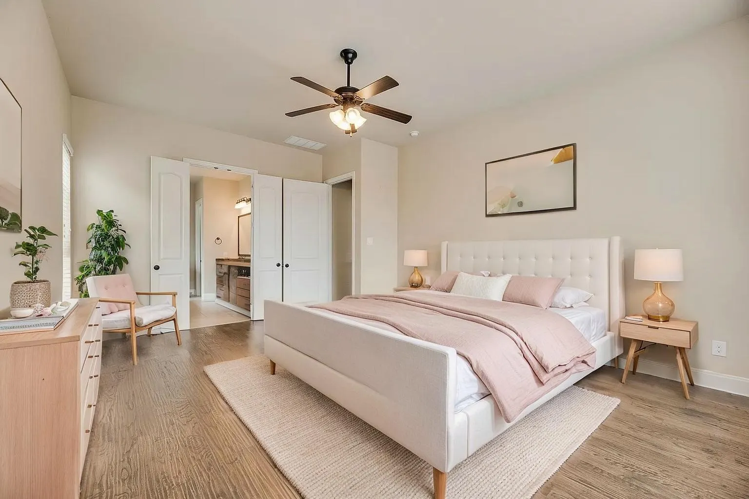 Wood-finish flooring throughout, ceiling fan with integrated lighting, and white double doors opening to a dual-vanity bathroom