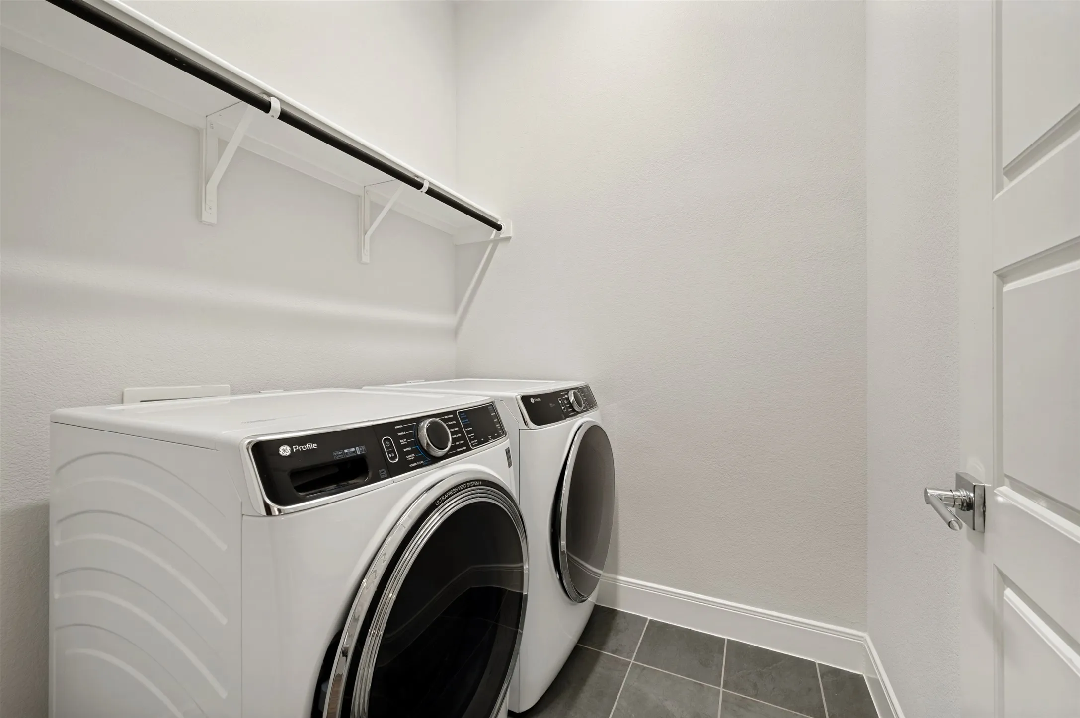 Laundry area featuring a front-load washer and dryer, white shelving with a hanging rod, and dark gray square tile flooring