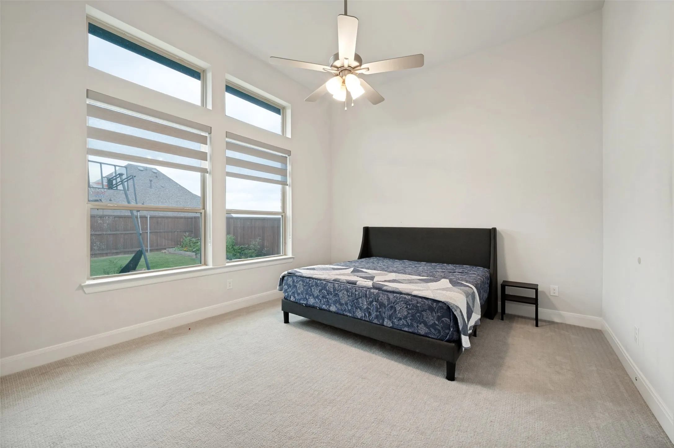 Carpeted bedroom featuring a large window with roller shades, a ceiling fan with integrated lighting, and high ceilings