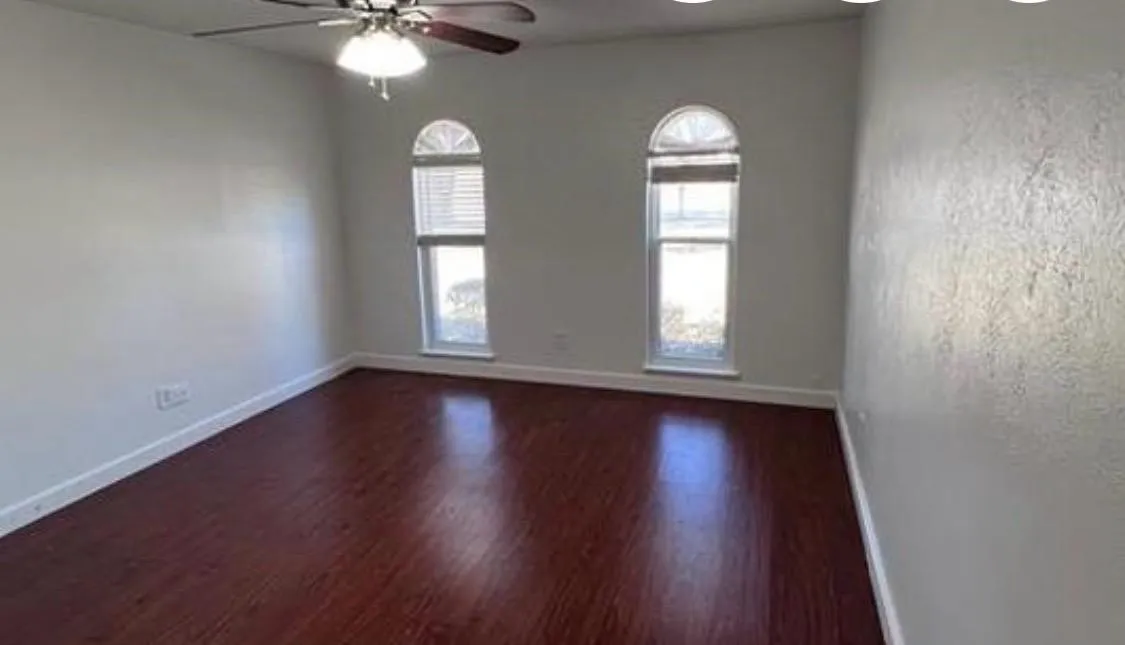 Room featuring wood-finish flooring, light-toned walls, two arched windows with blinds, and a ceiling fan with integrated lighting