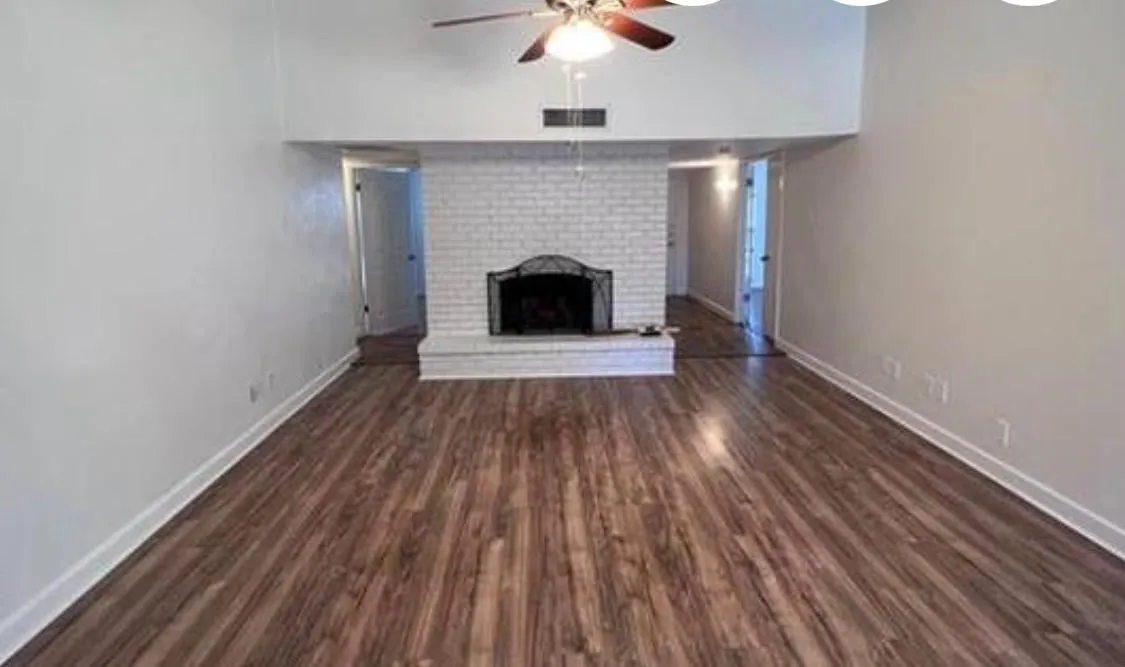 Spacious living area featuring wood-finish flooring, a white-painted brick fireplace with a hearth, and a ceiling fan
