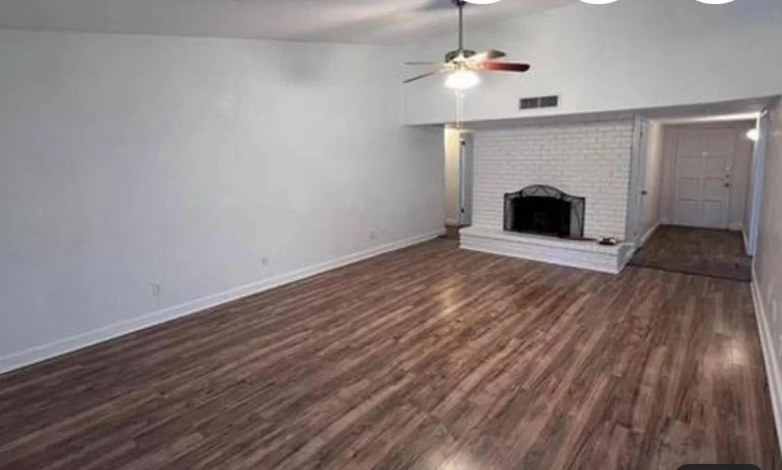 Spacious living area featuring wood-finish flooring, a white brick fireplace with a decorative metal screen, and a ceiling fan with integrated lighting