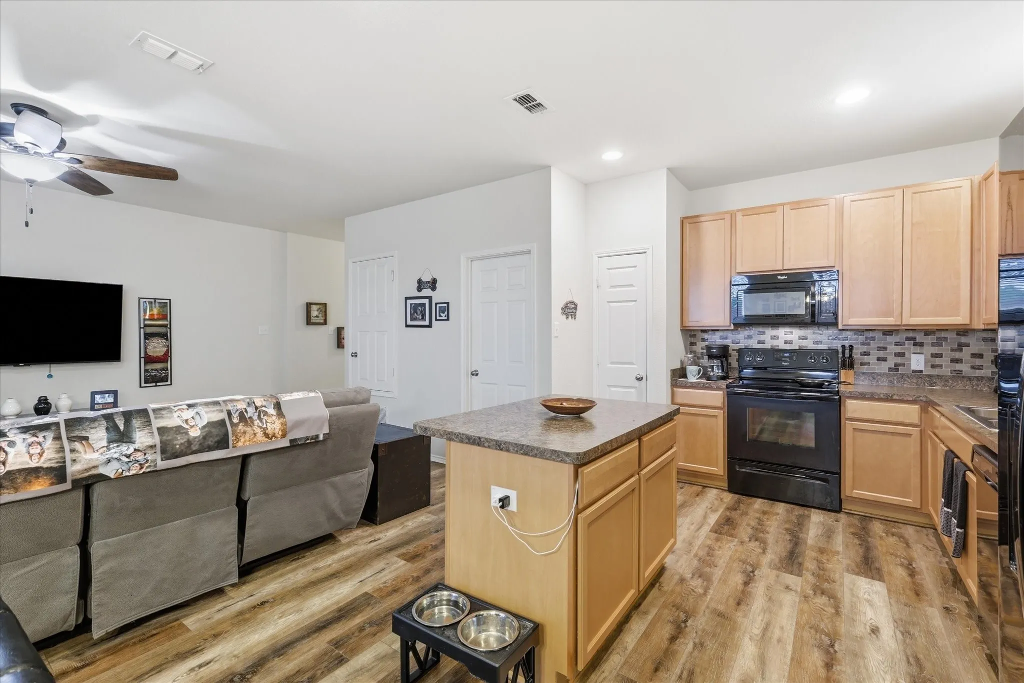 Open-concept living space featuring wood-finish flooring, a kitchen island with a stone-look countertop, light wood cabinetry, black appliances, and a tiled backsplash
