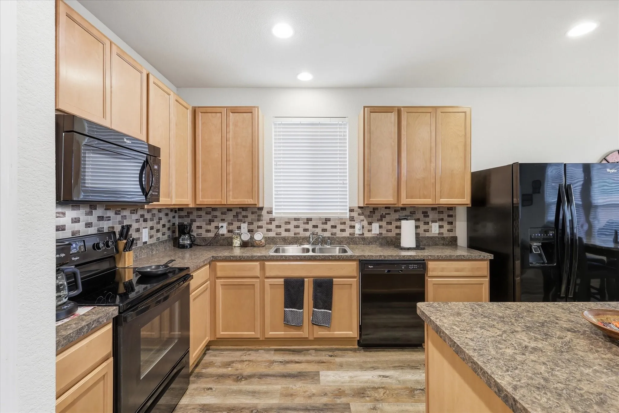 Kitchen featuring light wood-finish cabinetry, black appliance suite, mosaic tile backsplash, and wood-finish flooring